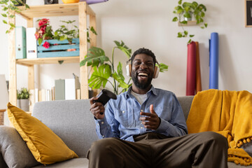 Cheerful non-binary person wearing wireless headphones and playing video game at home