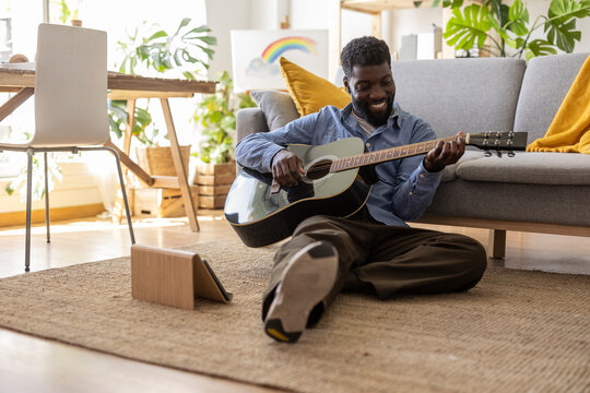 Happy non-binary person playing guitar near tablet PC at home