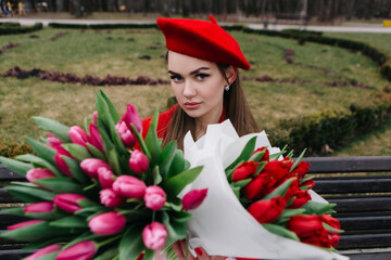 Young woman in red french beret sitting on bench cloudy weather in early spring nature with bouquet...