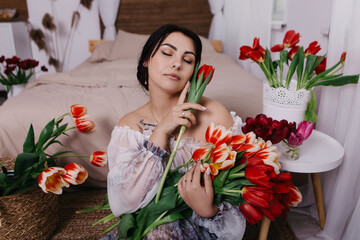 A woman poses near bed, surrounded by vibrant fresh bouquets bunch of red tulips placed around the room, domestic atmosphere. Spring International Women's Day, 8th of March. Holiday, festive 

