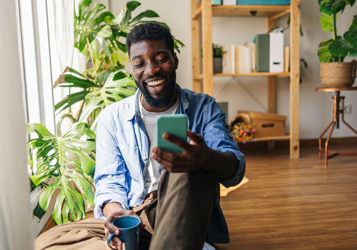 Cheerful non-binary person using smart phone and sitting with coffee cup at home