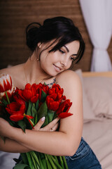 A woman poses on bed, surrounded by vibrant fresh bouquets bunch of red tulips placed around the room, domestic atmosphere. Spring International Women's Day, 8th of March. Holiday, festive 
