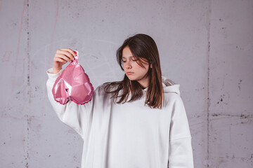 Girl holding deflated heart shaped pink balloon in front of cement background