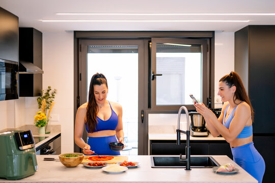 Woman photographing sister preparing pizza in kitchen at home