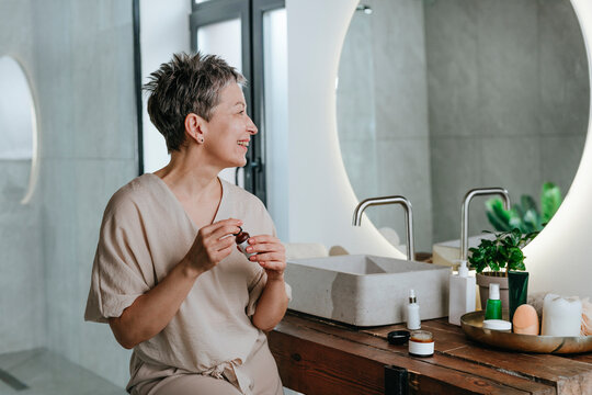 Smiling woman holding serum bottle near mirror at home