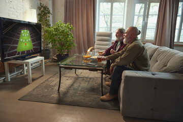 Elderly men, friends sitting on sofa in living room, eating snacks, drinking beer and watching...