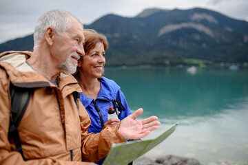 Portrait of beautiful active elderly couple hiking together in spring mountains. Senior tourists looking at map in front of lake.