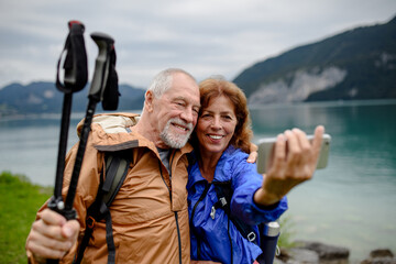 Portrait of beautiful active elderly couple taking selfie during hike in autumn mountains. Senior tourists embracing each other in front of lake - obrazy, fototapety, plakaty