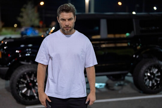 Angry man talking on phone near car on night urban street. Dangerous aggressive man talking on phone with serious face. Criminal city. Danger district. Aggressive angry man talking on phone outdoor.