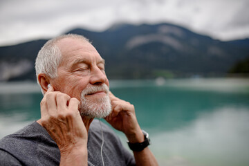 Senior listening music while running by lake in nature. Elderly man exercising to stay healthy, vital, enjoying physical activity and relaxation outdoors. - obrazy, fototapety, plakaty