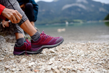 Close up of active woman putting on hiking boots on shore of mountain lake. Senior tourists enjoying nature during early spring.