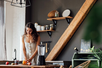 Charming curly-haired brunette girl in linen sundress prepares fruit smoothie in blender in kitchen