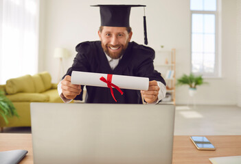 Happy graduate man showing his diploma via laptop having online video call. Male student sitting at...