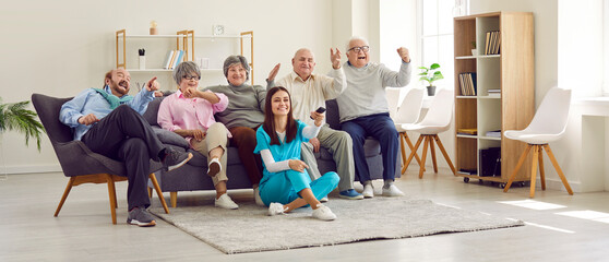 Group of senior people watching TV in retirement home together with young nurse. Active cheerful...