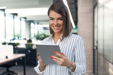 Portrait of young businesswoman looking at camera, holding touchpad while standing in modern office space interior. - 779506656