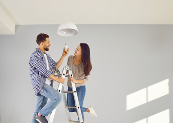 Cheerful young couple changing light bulb in apartment. Happy husband and wife standing on stairway...