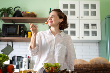 Young cheerful woman eating fresh vegetable salad while standing in the kitchen at home, healthy eating concept - 779521805