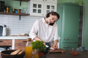 Young cheerful young woman talking on mobile phone while cooking at the kitchen. - 779522016