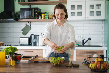 Young Caucasian woman cooking salad from green fresh vegetables while standing in the kitchen at...