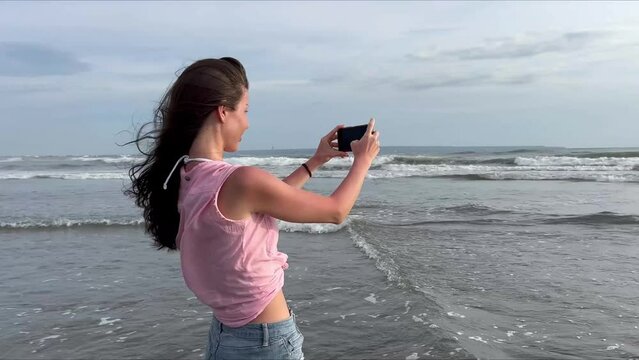 A beautiful girl takes pictures of the ocean on her smartphone. A woman walks along the coast of a popular beach and shoots a video of foaming sea waves at sunset for social media. . High quality 4k 