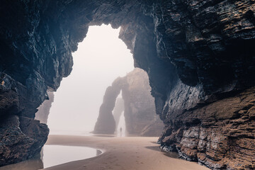 Man standing under natural arch on Cathedrals beach in Galicia, Spainn. Tourist silhouette in foggy landscape with Playa de Las Catedrales Catedrais beach in Ribadeo, Lugo on Cantabrian coast