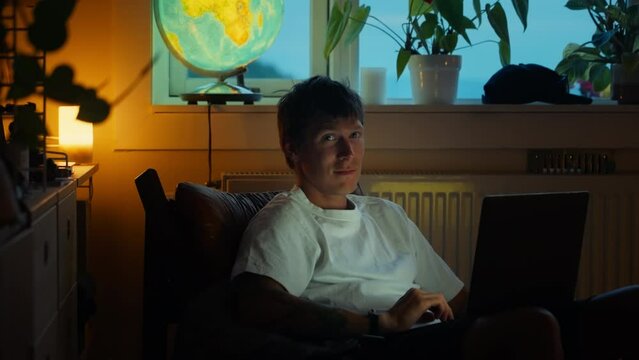 Portrait shot of young adult man look at laptop, screen lights up his face in darkness. Man at home work on project after hours or watch TV show, look at camera and smile.Cosy and safe home atmosphere