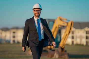 Successful construction business owner. Construction worker in suit and helmet near excavator....