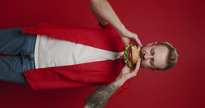 Vertical video of Unshaven caucasian man eats meat burger with pleasure on isolated red background, bites burger with his mouth, man casually eats junk food from fast food. Hungry Man Eat big burger.