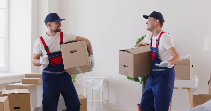 Two cheerful male movers have fun dancing with cardboard boxes during moving process. Loaders in uniform are having fun among cardboard boxes. Concept of moving services and easy working atmosphere.