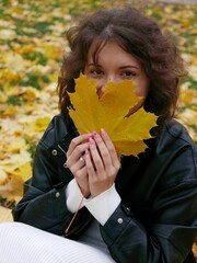 Vertical of curly brunette Caucasian female in black leather jacket posing with yellow maple leaves