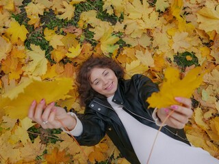 Top view of a curly brunette Caucasian female playing with yellow maple leaves on the ground