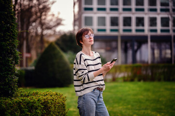 A thoughtful woman holding a smartphone stands amidst urban greenery, her upward gaze reflecting a moment of introspection in the city.