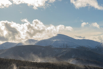 The sun shines through the clouds and snow dust against the backdrop of huge mountains.