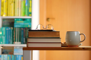 Cup of tea or coffee, pile of books, plate of cookies, reading glasses, e-reader and pen on the table. Colorful rainbow bookshelf in the background. Selective focus.