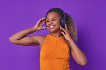 Young smiling beautiful African American woman meloman puts headphones on head to listen to music from own playlist or new musical album of pop group stands in purple studio. Podcast, audio book