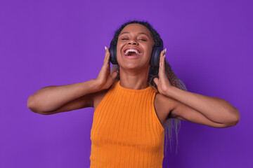 Young excited African American woman laughs pressing wireless headphones to ears listening to...
