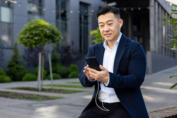 Asian young man in a business suit sitting outside on a bench near an office building wearing headphones and using a mobile phone