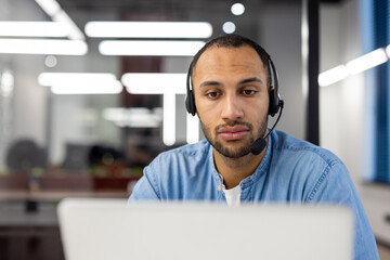 Concentrated and serious young hispanic man in headset working at laptop in office. Close-up photo