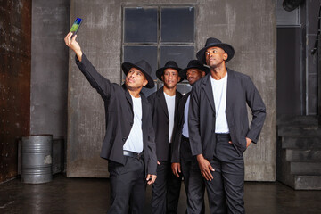 Group of male artists stands on stage in black suits with hats, exuding elegance and confidence in...