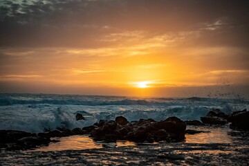 Fototapeta premium Beautiful golden sunset at a beach with waves hitting the silhouette of rocks on the shore