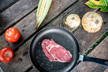 Top view of a black pan with seasoned raw steak on a wooden table with corn, tomatoes, and onions