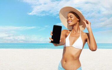 Happy young woman at the beach side showing mobile phone, wearing a turquoise sun hat and bikini, portrait of African latin American woman in sunny summer day with blue sky, concept of summer holiday