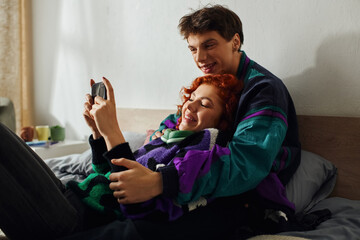 handsome joyous man with headphones using smartphone with his loving red haired girlfriend on bed
