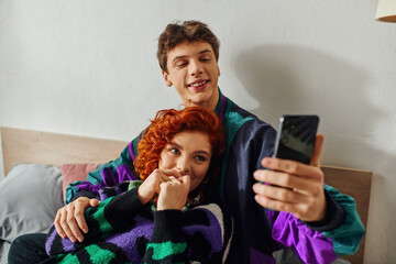 joyous young appealing couple in cozy attire looking at smartphone while lying on bed together
