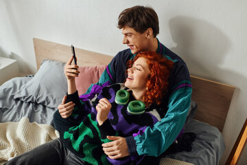 handsome cheerful man with modern headphones looking at phone with his joyous girlfriend on bed