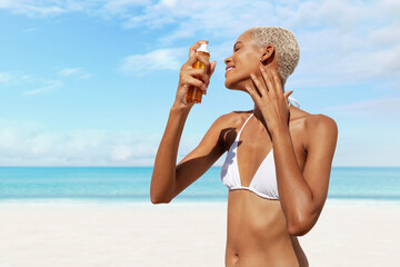 Woman at the beach shields herself from the sun applying sunscreen lotion on face in sunny day with blue sky. Concept of summer vacation, holiday travel, or sun protection for health and skincare.