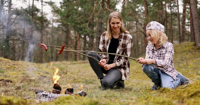mother and daughter frying sausages over a campfire in the forest. family time, bonding activities