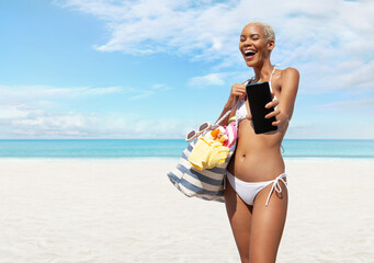 Happy woman at the beach side wearing bikini holding a beach bag and showing mobile phone in a sunny day with blue sky. Concept of summer beach holiday, shopping online, booking travel and resort
