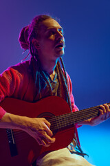 Portrait of young man, soulful musician with dreadlocks expressing himself through his guitar melodies against blue background in neon light. Concept of music, performance, festival, concert