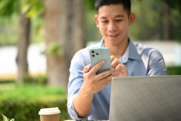 Smiling young businessman texting message on mobile phone sitting outside at cafe table